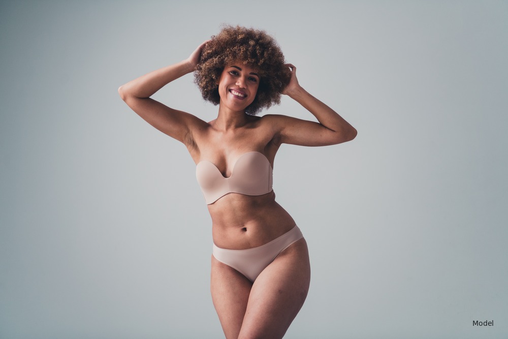 Woman with curly afro in nude lingerie, hands behind head, studio gray background